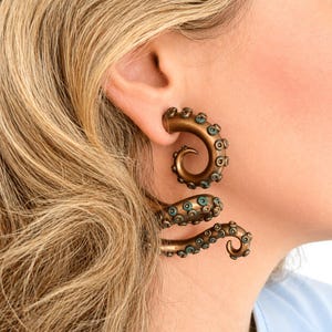 Puede incluir: Pendientes de bronce con forma de tentáculo de pulpo. Los pendientes presentan un diseño en espiral con pequeñas ventosas de color turquesa. Se llevan en la oreja y se extienden por el lóbulo, creando un look único. Miden aproximadamente 7 cm.