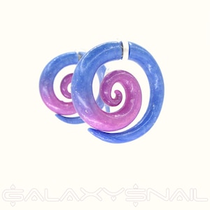 Puede incluir: Un par de pendientes en espiral con temática galáctica. Los pendientes presentan un degradado de tonos azules y morados, con un efecto moteado. El diseño es en espiral, con un cierre plateado. El fondo es blanco.