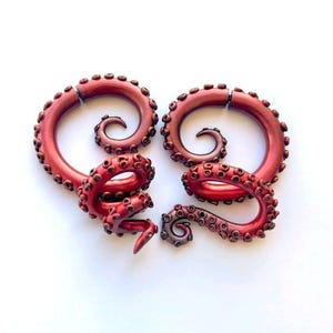 May include: A pair of dark red, spiral-shaped ear gauges designed to resemble octopus tentacles. Each piece features a curved, tapering form covered in raised, circular suction cup details along the outer edge, with a smooth, metallic finish.