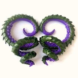 Peut inclure: Une paire d'écarteurs d'oreilles en forme de spirale avec une coque extérieure segmentée vert foncé et une surface intérieure violet vif couverte de petites ventouses circulaires en relief. Le design imite des tentacules recourbés.