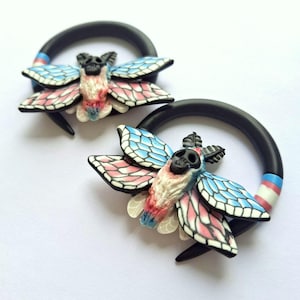 Puede incluir: Un par de dilatadores circulares negros con un adorno en forma de polilla. La polilla tiene una cabeza similar a una calavera, un cuerpo blanco y rosa, y alas con rayas azules, rosas y blancas. Una pequeña sección del aro muestra un diseño de rayas azules, rosas y blancas.
