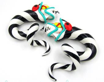 May include: A pair of whimsical, handmade polymer clay earrings. Each earring features a black and white striped body with a curled tail. The heads have turquoise accents, red eyes, and white teeth. The earrings are designed to resemble a cartoon snake.
