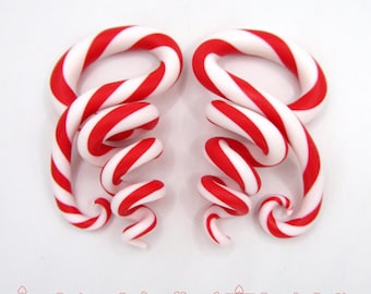 May include: A pair of red and white candy cane-shaped ear gauges. The gauges are made of polymer clay and are twisted into a spiral shape.