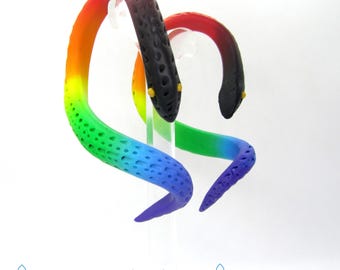 May include: A pair of rainbow-colored snake earrings with black spots. The earrings are made of polymer clay and are shaped like a snake with a curled tail. The earrings are displayed on a white background.