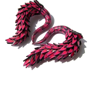 Peut inclure: Paire de jauges d'oreille décoratives roses et noires. Les jauges ont une forme incurvée et allongée avec un design en couches en forme de plume. Les bords sont soulignés en noir et la surface a une finition brillante.