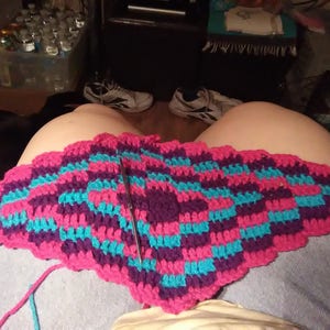 May include: A colorful, handmade crochet project in progress. The piece features a repeating pattern of pink, teal, and purple squares. A crochet hook is inserted into the work. The project is resting on a person's lap.