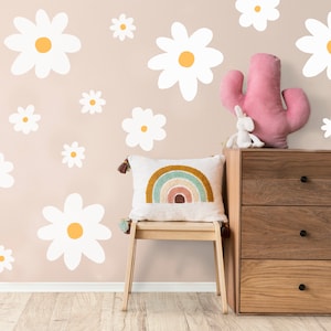 May include: A white wooden chair with a rainbow embroidered cushion sits in front of a chest of drawers with three drawers. The wall behind the chair is decorated with white daisy decals.