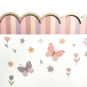 May include: A decorative wall panel with a scalloped wooden top and a striped background in shades of pink, purple, and beige. The lower section features a white surface adorned with pastel butterflies, flowers, and hearts.