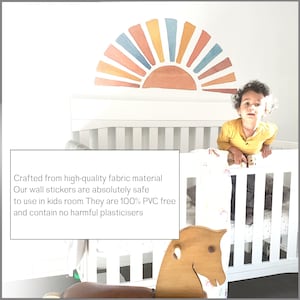 May include: A white crib with a rainbow-colored sun wall decal above it. A young child is standing in the crib, looking at the camera. The crib has a white rocking horse in front of it. The text "Crafted from high-quality fabric material Our wall stickers are absolutely safe to use in kids room. They are 100% PVC free and contain no harmful plasticisers" is visible in the image.