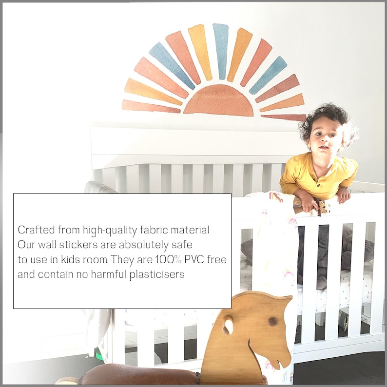 May include: A white crib with a rainbow-colored sun wall decal above it. A young child is standing in the crib, looking at the camera. The crib has a white rocking horse in front of it. The text "Crafted from high-quality fabric material Our wall stickers are absolutely safe to use in kids room. They are 100% PVC free and contain no harmful plasticisers" is visible in the image.