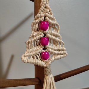 May include: A handmade macrame Christmas tree keychain. The tree is crafted from beige rope and adorned with three bright pink beads. A silver keyring is attached at the top, and a tassel hangs from the bottom. The tree is displayed against a wooden background.