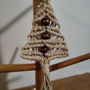May include: A handmade macrame Christmas tree keychain. The tree is crafted from natural-colored twine, featuring three dark wooden beads. The keychain is attached to a silver metal ring, with a tassel at the bottom. The tree is approximately 3 inches tall.