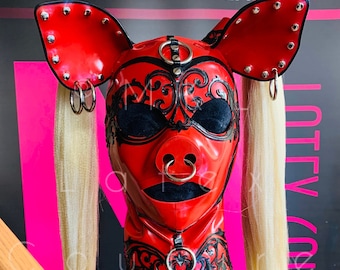 May include: A red latex hood with pig-like features, including large ears and a snout, adorned with silver studs and rings. The hood has black accents and long, blonde hair extensions. The background features the text "www.mrlatex.com".