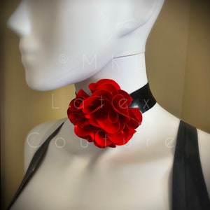 May include: A black choker necklace with a large, vibrant red flower accent. The flower has multiple layered petals. The necklace is displayed on a white mannequin. The image has a soft focus.