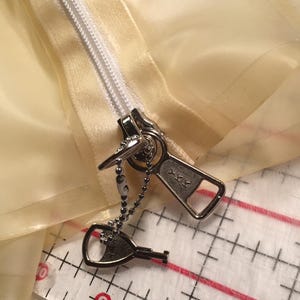 May include: Close-up of a silver zipper pull with a key-shaped charm attached by a beaded chain. The zipper is partially closed on a pale yellow fabric. The zipper pull is marked with the letters "YKK".