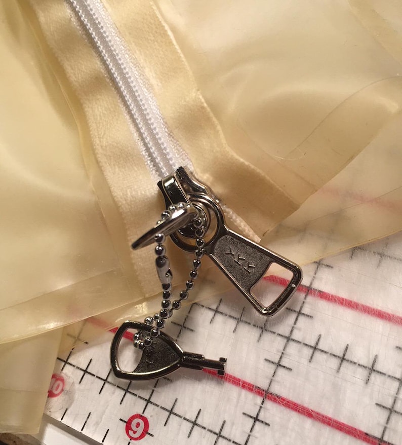 May include: Close-up of a silver zipper pull with a key-shaped charm attached by a beaded chain. The zipper is partially closed on a pale yellow fabric. The zipper pull is marked with the letters "YKK".