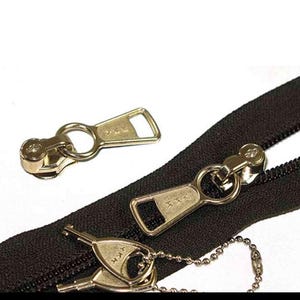 May include: Close-up of several gold-colored zipper pulls and a black zipper. The zipper pulls have a rectangular design with a hole at the top and the text "YKK" on them. One pull is attached to a chain.