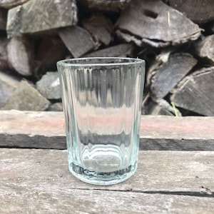 Puede incluir: Un vaso de chupito de vidrio transparente con un diseño acanalado. El vaso está vacío y se encuentra sobre una superficie de madera.