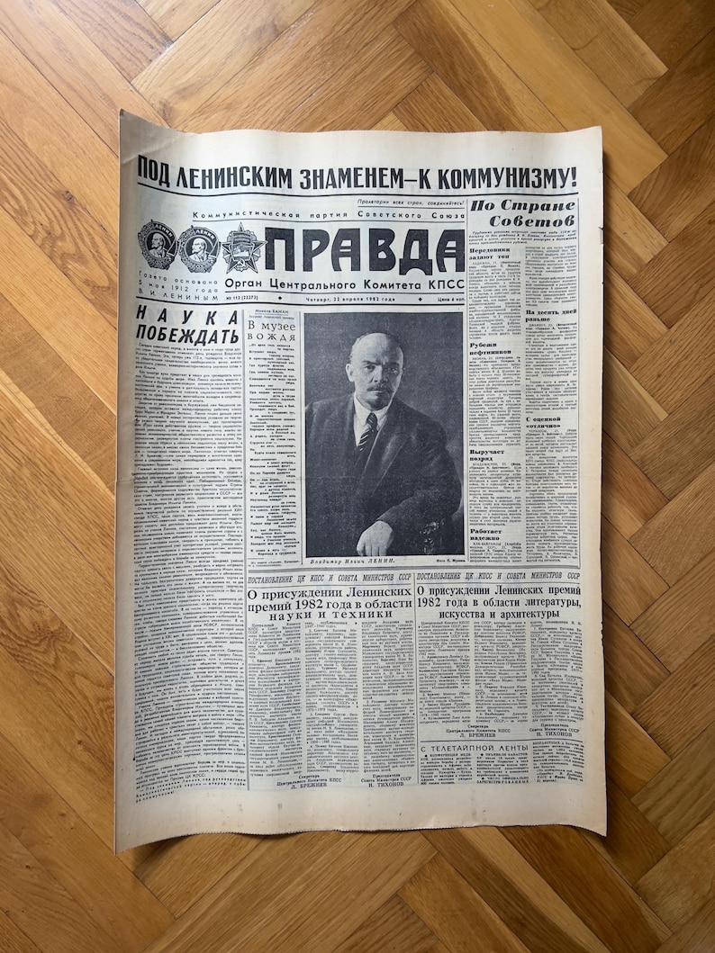May include: A black and white newspaper with the headline "POD LENINSKIM ZNAMENEM-K KOMMUNIZMU!" and the title "PRAVDA" in bold, red letters. The newspaper is dated "22 июня 1982 года" and features a portrait of Vladimir Lenin.