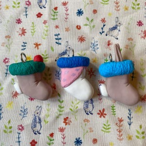 May include: Three handmade felt Christmas stockings with blue, green, and pink trim. The stockings are decorated with a red pom-pom, a pink and white patterned fabric, and a blue felt cuff.
