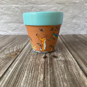 Könnte beinhalten: Ein Terrakotta-Pflanztopf mit einem hellblau bemalten Rand. Die Außenseite zeigt eine handgemalte Szene mit einem orangefarbenen Hasen auf einem grauen Stein, einem kleinen blauen Vogel auf einem grünen Zweig und schwarzen Insekten.