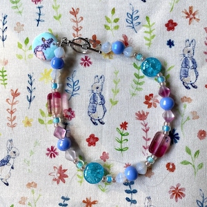 May include: A blue and pink beaded bracelet with a Peter Rabbit charm. The bracelet has a silver clasp and is made with glass beads.