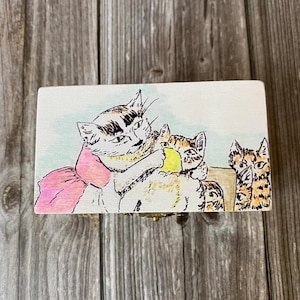 May include: A rectangular wooden box featuring a hand-drawn illustration of three cats. One cat wears a large pink bow, another holds a yellow ball, and a third cat sits to the right. The artwork uses black ink outlines with soft watercolor accents.
