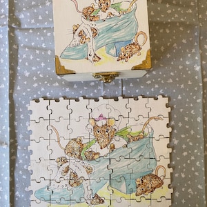 Op de afbeelding: Een witte houten doos met een scharnierend deksel en een messing sluiting. De doos is versierd met een handgeschilderde illustratie van een groep muizen in een blauwe schoen. De muizen dragen kleding en hebben menselijke gezichtsuitdrukkingen.