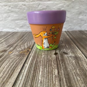 Könnte beinhalten: Ein Terrakotta-Pflanztopf mit einem handgemalten orangefarbenen Hasen, der einen bunten Blumenstrauß auf einem grünen Grasfleck hält. Der Rand des Topfes ist in einem einfarbigen Violett bemalt, das einen Kontrast zum natürlichen Ton bildet.