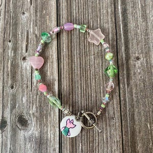 Puede incluir: Una pulsera de cuentas con una variedad de cuentas de vidrio rosas y verdes en formas que incluyen corazones, estrellas y esferas. La pulsera incluye un cierre de palanca plateado y un dije circular que representa un cerdo rosa.