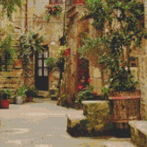 May include: A view of a narrow, cobblestone street in a European village. The street is lined with buildings with terracotta roofs and lush green foliage. The buildings have windows and doors, and there are potted plants and flowers throughout the scene.