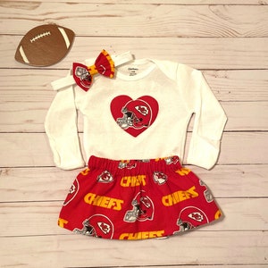 May include: A white long-sleeve onesie with a red heart-shaped design featuring a football helmet. A red skirt with a Chiefs helmet pattern and a matching bow headband complete the set. A football-shaped decoration is also present.