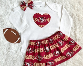 May include: A white long-sleeve bodysuit featuring a red heart with the San Francisco 49ers logo, a red skirt with a repeating 49ers logo and text pattern, a matching red and white hair bow, and a brown football-shaped cutout.