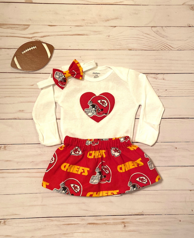 May include: A white long-sleeve onesie with a red heart-shaped design featuring a football helmet. A red skirt with a Chiefs helmet pattern and a matching bow headband complete the set. A football-shaped decoration is also present.