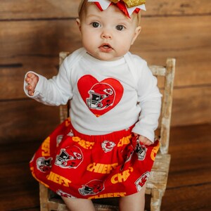 May include: A baby wearing a white long-sleeved shirt with a red heart and football helmet design, paired with a red skirt featuring football helmets and the word "CHIEFS". A large red, white, and gold bow adorns the baby's head.