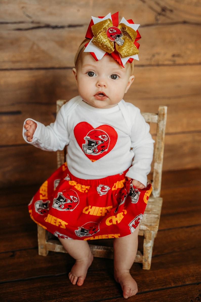 May include: A baby wearing a white long-sleeved shirt with a red heart and football helmet design, paired with a red skirt featuring football helmets and the word "CHIEFS". A large red, white, and gold bow adorns the baby's head.