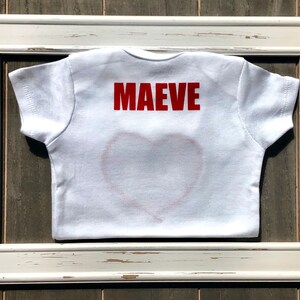 May include: White baby t-shirt with the name "MAEVE" in red block letters on the back. A red heart outline is visible on the front. The shirt is framed in a distressed white wooden frame.