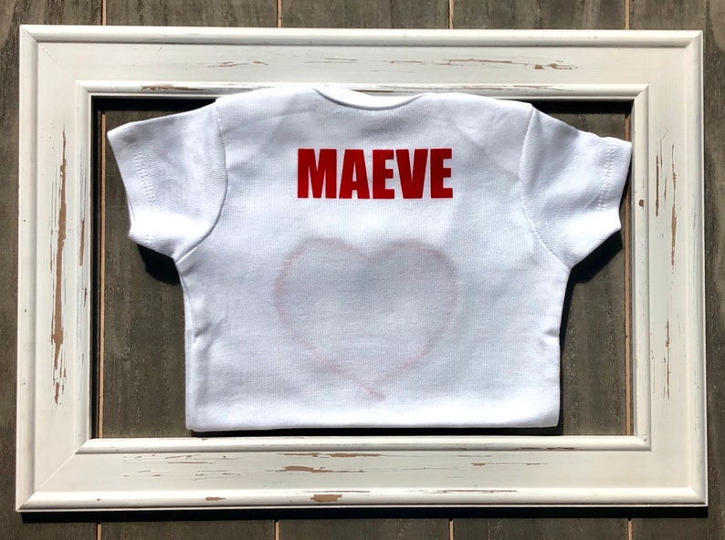 May include: White baby t-shirt with the name "MAEVE" in red block letters on the back. A red heart outline is visible on the front. The shirt is framed in a distressed white wooden frame.