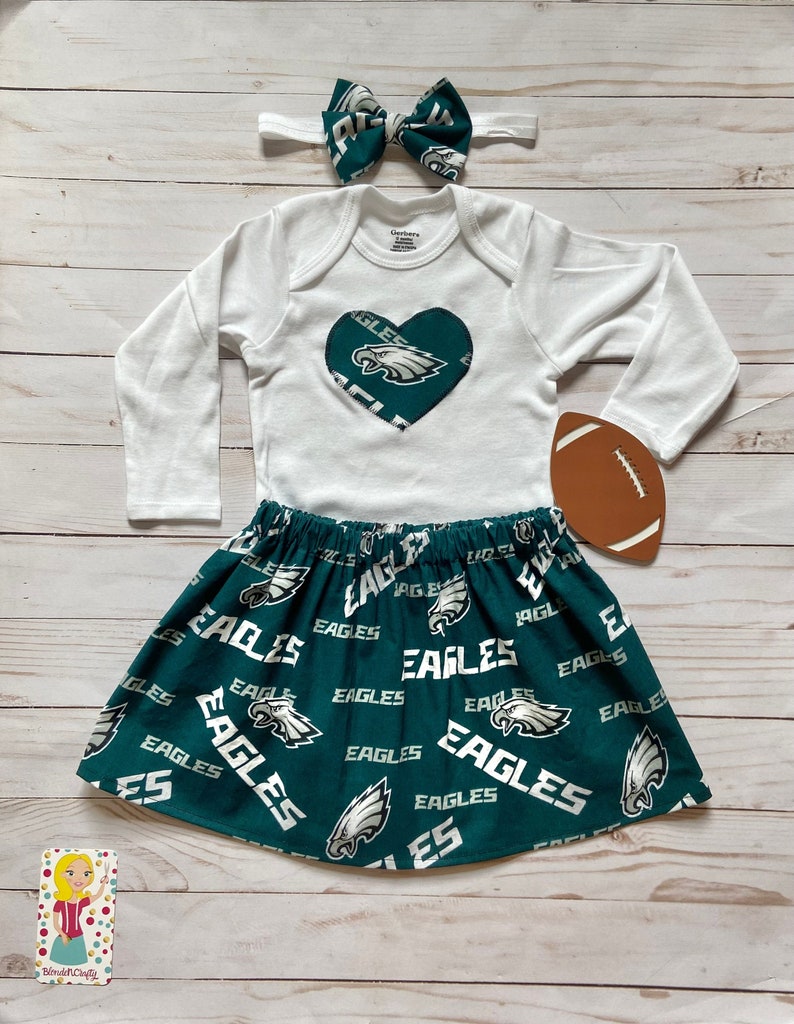 Peut inclure: Une tenue de b&eacute;b&eacute; avec un body blanc &agrave; manches longues orn&eacute; d'un c&oelig;ur vert avec un logo d'aigle blanc. La tenue comprend &eacute;galement une jupe verte avec un logo d'aigle blanc et le mot "Eagles" r&eacute;p&eacute;t&eacute; partout. Un bandeau vert et blanc et un ballon de football marron sont &eacute;galement inclus.