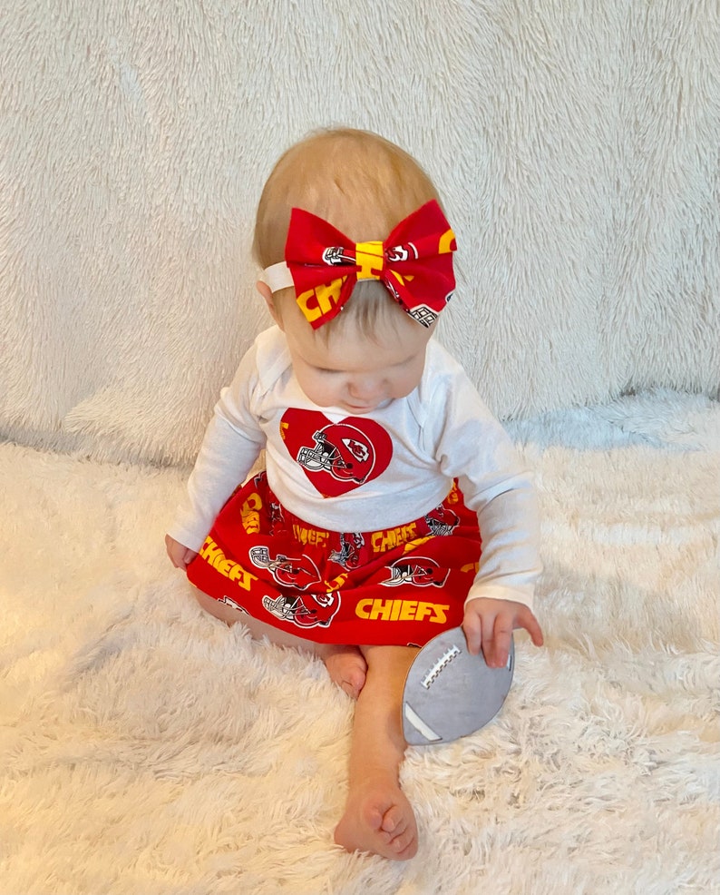 May include: A baby wearing a red and yellow Kansas City Chiefs themed outfit. The outfit includes a white long-sleeved shirt with a heart-shaped logo, a red skirt, and a matching bow headband. A gray football-shaped toy is held in the baby's hand.