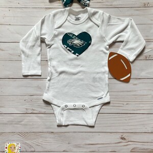 Peut inclure: Combinaison blanche pour b&eacute;b&eacute; avec un c&oelig;ur vert orn&eacute; du logo des Philadelphia Eagles. Un bandeau avec un n&oelig;ud papillon vert et blanc et un ballon de football marron sont &eacute;galement inclus.