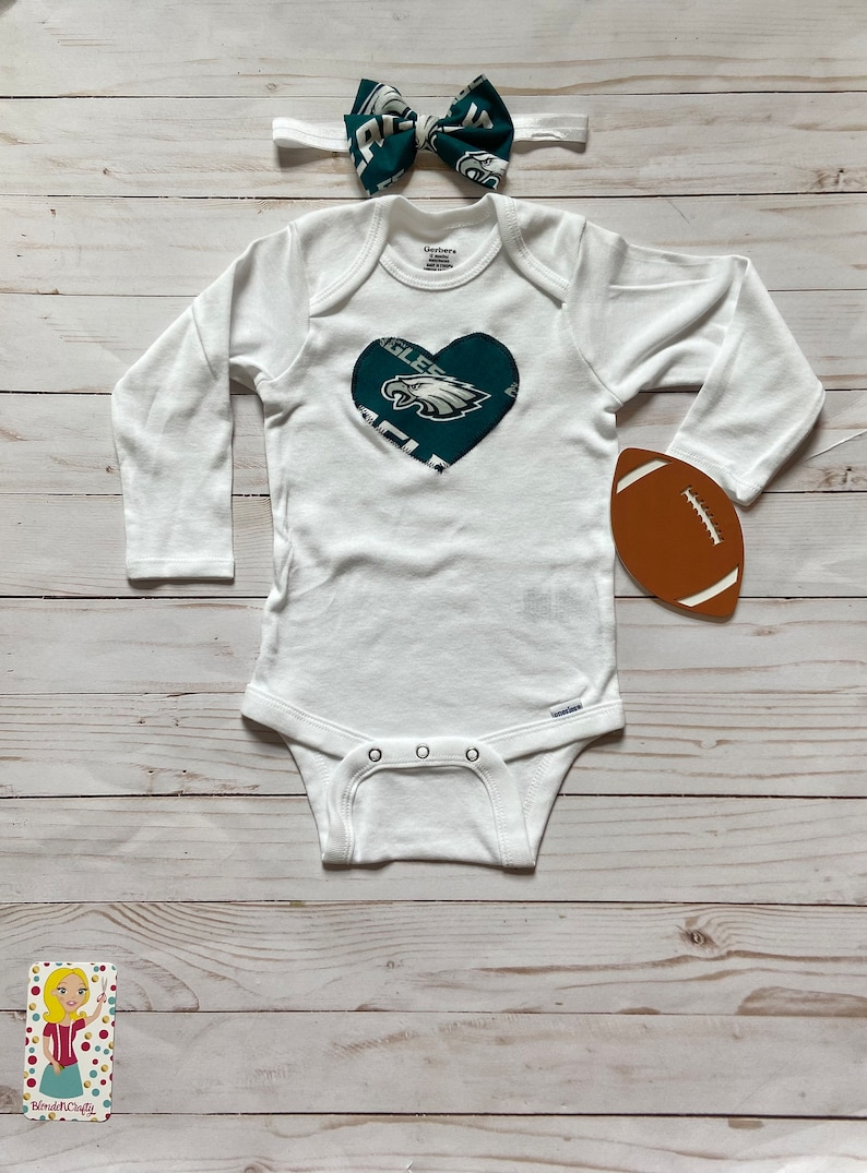Peut inclure: Combinaison blanche pour b&eacute;b&eacute; avec un c&oelig;ur vert orn&eacute; du logo des Philadelphia Eagles. Un bandeau avec un n&oelig;ud papillon vert et blanc et un ballon de football marron sont &eacute;galement inclus.