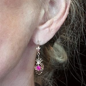May include: A gold-colored earring with a pink gemstone. The earring has a decorative design with a crown-like top and a circular element holding the gemstone. The earring is hanging near a person's ear.