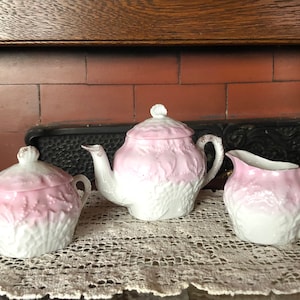 May include: A vintage tea set with a pink and white floral design. The set includes a teapot, creamer, and sugar bowl.
