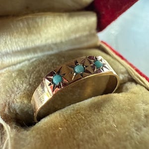 May include: A gold band ring featuring three small, round, light blue turquoise stones set within individual star-shaped engravings. The ring is displayed inside a red velvet-lined jewelry box with a cream-colored fabric interior.
