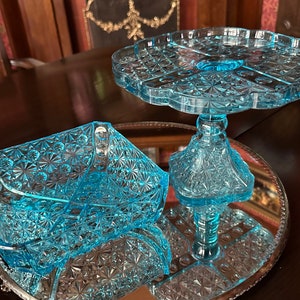 May include: A set of two blue glass serving dishes with a cut glass pattern. The larger dish is a pedestal cake stand and the smaller dish is a square bowl with feet.