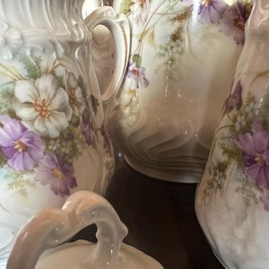 Peut inclure: Gros plan de plusieurs pichets en céramique blanche avec des motifs floraux. Les pichets sont décorés de fleurs violettes et blanches, de feuilles vertes et de détails délicats. Les pichets ont des anses ornées et une finition brillante.