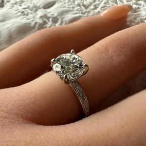 May include: A sparkling diamond ring with a large, round-cut center stone. The ring is set in a silver band, with smaller diamonds accenting the band. The ring is shown on a person's finger.