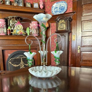 May include: A multi-tiered glass epergne featuring a central trumpet-shaped vase and two smaller hanging flower-shaped vases, all with white opalescent glass, ruffled edges, and decorative green glass vines, set on a ruffled glass base.