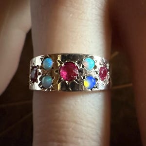 May include: A gold ring with alternating red and iridescent blue gemstones. The ring's band has a decorative, openwork design. The gemstones are round and set into the band. The ring is on a finger.
